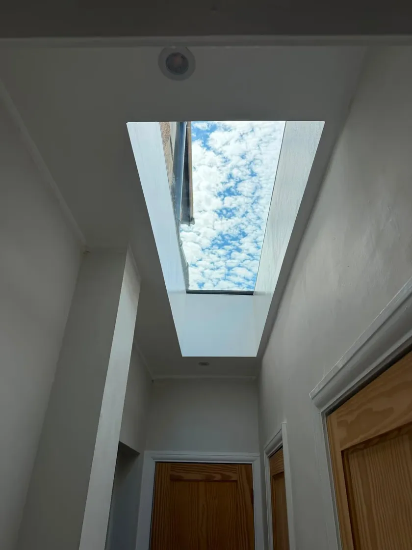 Looking up through a flat glass rooflight and splayed white light shaft to blue sky and cloud, above a hallway in Worcester Park