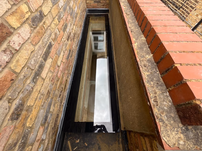 Newly installed flat glass rooflight seen from roof level, sitting in the narrow gap beside the neighbouring brick wall