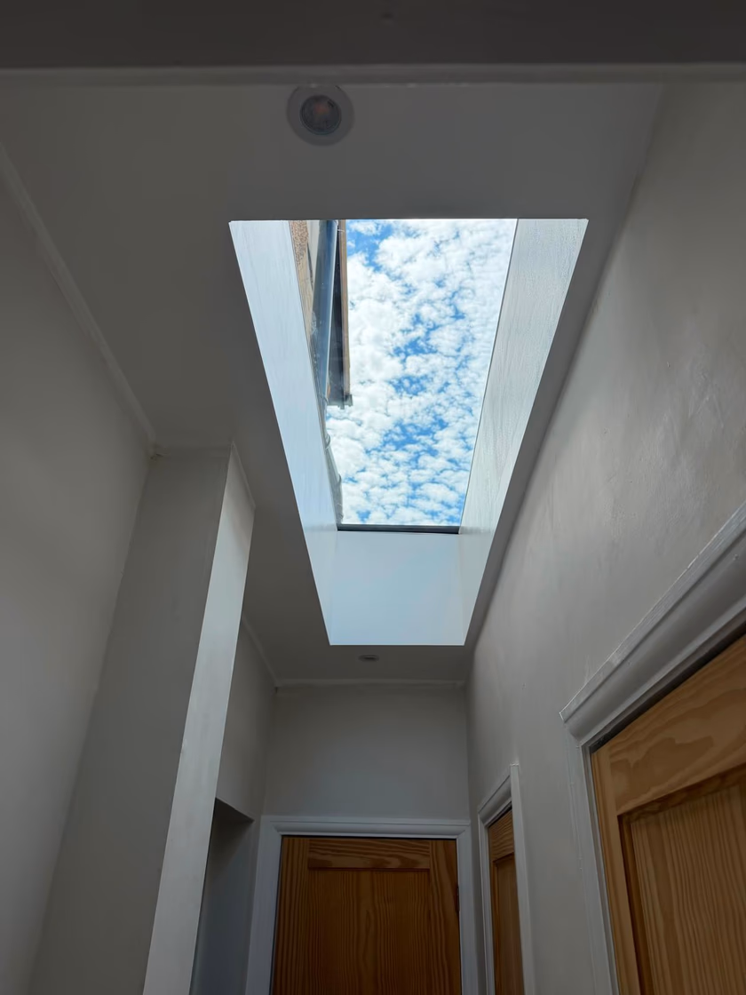 Finished flat glass rooflight above a hallway, with a splayed white plastered shaft opening to blue sky and cloud
